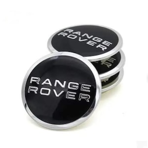 May include: A stack of four black and silver Range Rover emblem badges. Each badge is round with the words "RANGE ROVER" in silver lettering against a black background. The badges have a shiny, reflective surface.