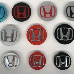 May include: A collection of various Honda wheel center caps. The caps are round and feature the Honda logo in different colors, including black, red, silver, and blue. The logos are in the center of the caps.