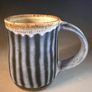 May include: A ceramic mug featuring a vertical striped pattern in dark green and light blue. The rim has a speckled brown and cream glaze that drips down onto the stripes. The mug includes a curved handle with a speckled grey and blue finish.