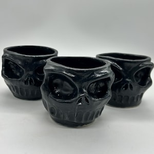 Puede incluir: Tres recipientes negros de cerámica con forma de calavera. Cada recipiente tiene una abertura ancha y está diseñado para parecerse a un cráneo humano. Los recipientes son brillantes y se asientan sobre una superficie blanca.