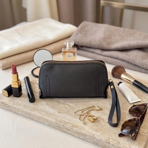 May include: A black rectangular zippered pouch with a wrist strap, surrounded by a lipstick, a compact mirror, a perfume bottle, a makeup brush, a tube of cream, a gold chain necklace, a gold ring, and brown tortoiseshell sunglasses.