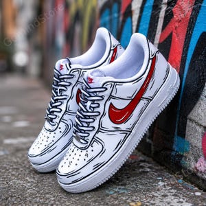 Peut inclure: Une paire de baskets basses blanches au design inspiré des bandes dessinées avec des contours noirs marqués, un swoosh Nike rouge sur le côté et le logo Nike rouge sur la languette. Les chaussures ont des lacets blancs et une semelle épaisse.
