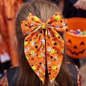 Könnte beinhalten: Eine große orangefarbene Haarschleife mit einem wiederkehrenden Halloween-Muster aus weißen Geistern, schwarzen Spinnennetzen, gelben Monden und orangefarbenen Kürbissen. Die Schleife hat zwei lange, spitz zulaufende Enden und einen Knoten.