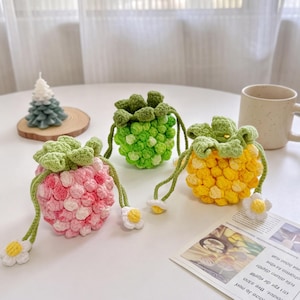 May include: Three small, round, crocheted drawstring pouches shaped like berries. One is pink, one is light green, and one is yellow. Each pouch features a textured, bumpy surface, a green crocheted leaf top, and drawstrings with small white flowers.