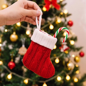 May include: A small red knitted Christmas stocking with a white ribbed cuff and a white hanging loop. A red, white, and green striped candy cane is tucked inside the top of the stocking, which features a vertical cable knit pattern.