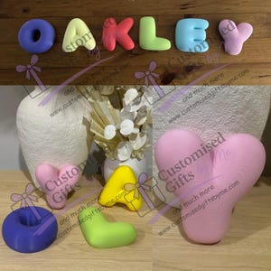 May include: A collection of 3D printed letters spelling OAKLEY in various pastel colors including purple, yellow, red, green, blue, and pink. The letters have a smooth, rounded, bubble-like appearance and are displayed on a wooden surface.