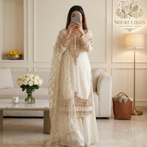 May include: Cream-coloured ethnic wear ensemble with intricate gold embroidery. The outfit includes a long-sleeved tunic, wide-leg trousers, and a sheer scarf. The design features detailed patterns on the neckline, sleeves, and hem.
