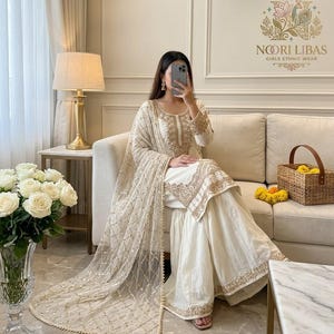 May include: A cream-coloured ethnic outfit with gold embroidery. The ensemble comprises a long-sleeved top, wide-legged trousers, and a sheer scarf. The outfit is worn with sandals. The image also shows a sofa, flowers, and a basket.