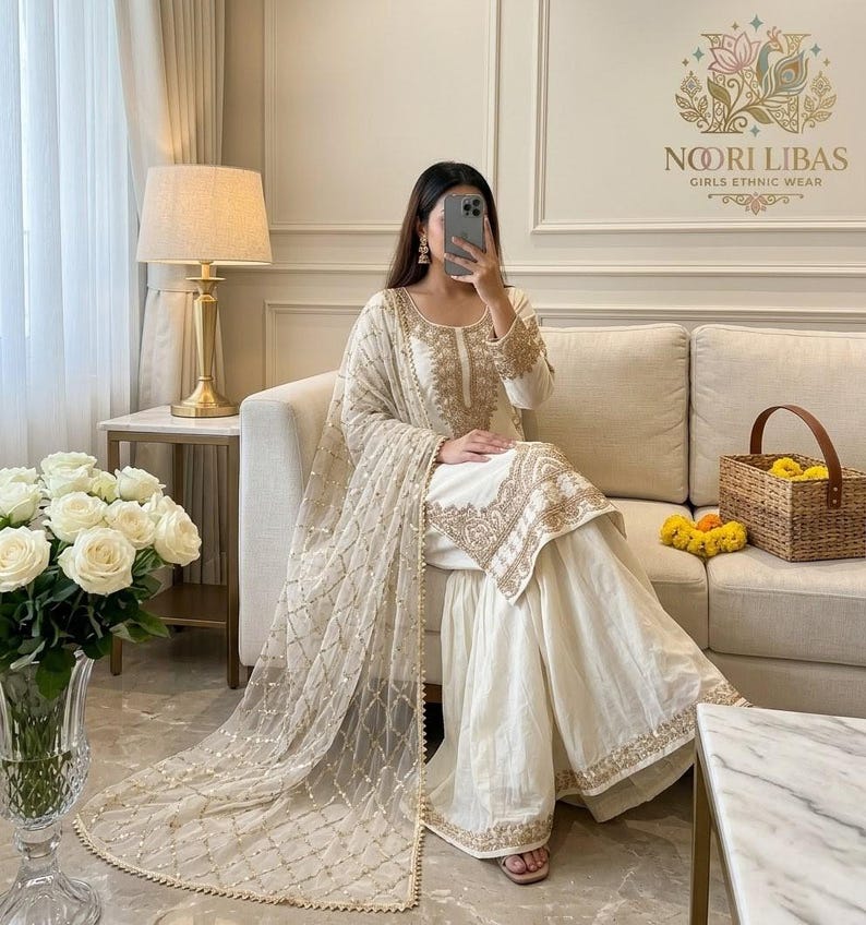 May include: A cream-coloured ethnic outfit with gold embroidery. The ensemble comprises a long-sleeved top, wide-legged trousers, and a sheer scarf. The outfit is worn with sandals. The image also shows a sofa, flowers, and a basket.