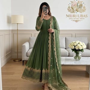 May include: An olive green ethnic wear dress with gold embroidery on the hem and sleeves. The dress features a flowing silhouette and is paired with a matching sheer dupatta. The dress has a v-neckline and button details.