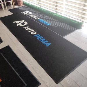 May include: Black rectangular floor mats with the text "AP AUTO PEMA" in white and blue. The mats are made of a textured material and are placed on a light-colored wooden floor. A rolled-up green mat is visible in the background.