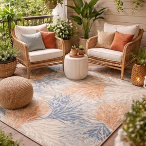 May include: An outdoor patio setting with two wicker armchairs, cream cushions, and orange and blue accent pillows. A patterned rug with blue and orange leaf designs covers the floor. Plants and string lights add to the ambiance.