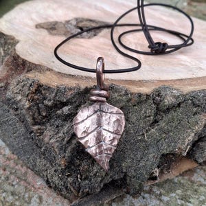 May include: A copper leaf pendant with a textured surface and a dark cord. The leaf-shaped pendant has visible veins and is suspended from a black cord. The pendant is made of copper and has a rustic, handmade appearance.