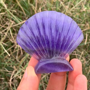 May include: A translucent purple carved stone figurine shaped like a seashell. The object features a fan-like design with vertical ridges radiating from a solid base, displaying deep violet hues and natural internal variations within the material.