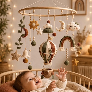 Könnte beinhalten: Handgefertigtes Baby-Mobile mit Holzring und gehäkelten Elementen. Das Mobile zeigt einen Heißluftballon, Sonne, Sterne, Mond, Wolke und Regenbogen in Grün-, Braun- und Cremetönen. Das Baby liegt in einem Holzbettchen.