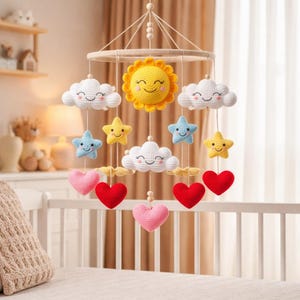 May include: A handmade baby mobile featuring a smiling yellow sun, white clouds, blue and yellow stars, and red and pink hearts. The mobile is suspended from a wooden ring and is designed to hang over a crib.