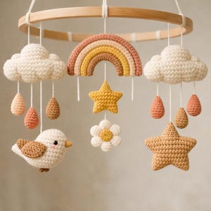 May include: A crochet mobile featuring a circular wooden frame with hanging elements including two white clouds with dangling teardrops, a central rainbow, a yellow star, a white flower, a small bird, and a large textured tan star.