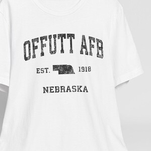 May include: White t-shirt with the text "OFFUTT AFB" in distressed black lettering. Below the text are the words "EST. 1918 NEBRASKA". A white baseball cap rests on a wooden chair behind the shirt.
