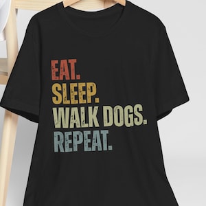 May include: Black t-shirt with the text "EAT. SLEEP. WALK DOGS. REPEAT." in a retro font. The text is in red, yellow, and blue-green. A white baseball cap rests on a wooden chair behind the shirt.