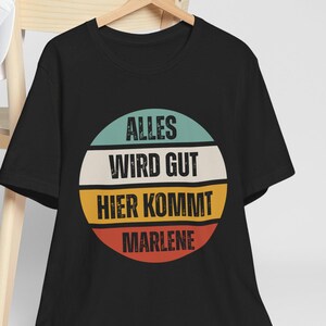 May include: Black t-shirt with a circular design featuring the text "ALLES WIRD GUT HIER KOMMT MARLENE" in white, teal, yellow, and red. The shirt is hanging on a wooden hanger. A white baseball cap rests on a wooden chair.