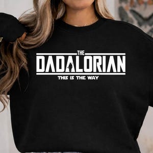 以下が含まれることがあります： 胸元に白い文字でTHE DADALORIANとプリントされた黒の長袖スウェットシャツ。Aの文字の中央には人物のシルエットが描かれ、その下に太字のサンセリフ体でTHIS IS THE WAYというテキストが配置されています。