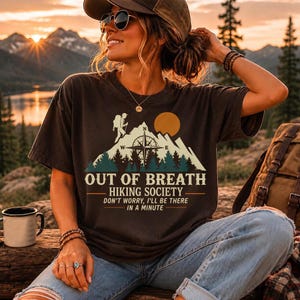 May include: A dark brown short-sleeve t-shirt featuring a graphic print of a mountain landscape, a compass, and a hiker. The text reads: OUT OF BREATH HIKING SOCIETY, DON'T WORRY, I'LL BE THERE IN A MINUTE. The design includes a sun and pine trees.