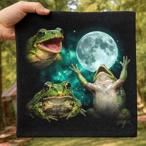 May include: A black square towel featuring a graphic print of three green frogs. One frog has an open mouth, another sits looking forward, and the third reaches upward toward a large, glowing full moon set against a teal, starry nebula background.
