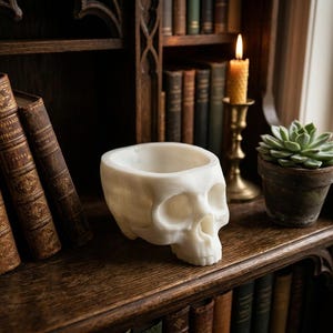 May include: A white, hollowed-out skull sculpture shaped into a bowl. The object features realistic anatomical details including eye sockets, a nasal cavity, and teeth. The smooth, matte-textured surface is carved to create a functional container.