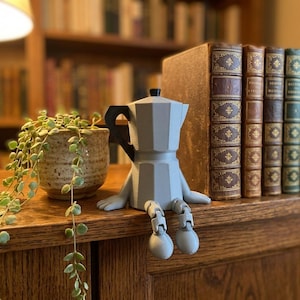 May include: A light gray, faceted moka pot figurine with a black handle, articulated arms, and jointed legs that hang over the edge. The coffee maker character features a geometric design and is positioned upright on a flat surface.