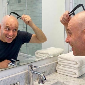 May include: A black plastic grooming tool with a curved handle and fine teeth, featuring the white text "BALD LAB" printed on the side, held against a smooth, hairless scalp while reflected in a bathroom mirror above a marble vanity.