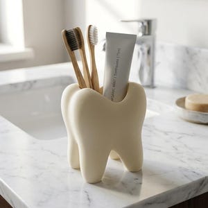 May include: A cream-colored, tooth-shaped holder containing two bamboo toothbrushes and a tube of toothpaste labeled Organic Mint | Toothpaste | 75ml. The holder has a smooth, matte finish and stands on four root-like legs, providing a stable base.