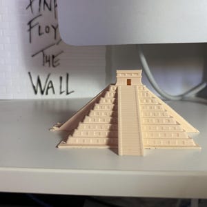 May include: A small, beige 3D-printed model of the Mayan pyramid El Castillo. The pyramid has a stepped design with a central staircase and a small temple at the top. The background includes a white brick wall with the words "PINK FLOYD THE WALL" written on it.