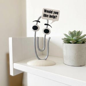 May include: A gray paperclip figurine with large white eyes, black eyebrows, and a white speech bubble sign that reads Would you like help? The figurine stands on a circular base next to a small green succulent in a gray concrete pot.