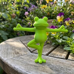 May include: A bright lime green frog figurine standing on one leg in a yoga pose with arms extended outward and fingers in a mudra position. The frog has a smooth surface, a wide mouth, and two small eyes on top of its head, balanced on a flat surface.