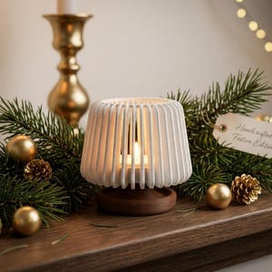 May include: A white, ribbed, cylindrical candle holder with a vertical slat design sits on a dark brown circular base. A small, lit candle is visible inside. A white paper tag attached to the side features the text Handcrafted - Festive Edition.