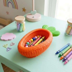 May include: An orange carrot-shaped storage bowl with a textured exterior and green leafy top, containing colorful beads, paper clips, and a yellow crayon. Nearby are several crayons, heart-shaped paper clips, and a pink cloud-shaped notepad.