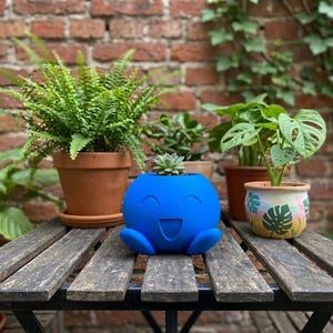 May include: A bright blue spherical planter featuring a carved smiling face with wide eyes and an open mouth. The planter contains a small green succulent plant and is positioned on a wooden slatted surface among several other potted plants.