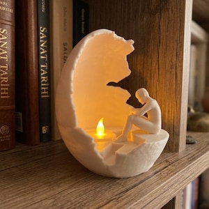 May include: A white, egg-shaped sculpture featuring a hollow interior with a seated, faceless figure. The sculpture contains a small, battery-operated LED tea light that emits a warm, glowing yellow flame, illuminating the interior space.