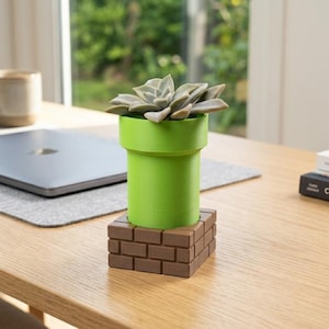 May include: A green cylindrical planter with a wide rim, designed to resemble a pipe, sits atop a brown square base with a brick pattern. A succulent plant with thick, fleshy, light green leaves is planted inside the green pipe-shaped container.