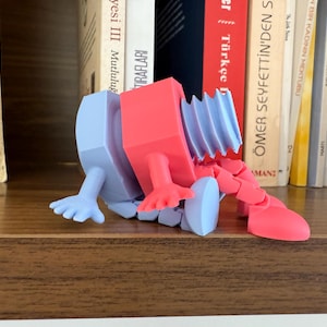 May include: A 3D-printed articulated toy in blue and red. The toy has a rectangular body, a flexible neck, and jointed limbs. The toy is resting on a wooden shelf in front of a row of books.