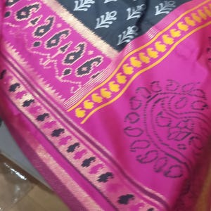 May include: A black fabric textile featuring a repeating white geometric pattern across the main body, bordered by a wide, vibrant pink section. This pink border displays intricate black paisley motifs and a thin, shimmering gold metallic stripe.