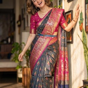 May include: A charcoal gray saree featuring a vibrant magenta border with intricate gold zari peacock motifs and floral patterns. The matching magenta blouse has vertical stripes and gold accents. A thin blue belt is worn around the waist.