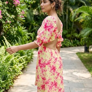 Könnte beinhalten: Gelbes Kleid mit rosa Blumenmuster. Das Kleid hat kurze, gepuffte Ärmel und einen Rückenausschnitt. Die Person befindet sich in einem Garten.