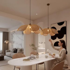 May include: Two flower-shaped pendant lights made of woven material hang above a white oval dining table. The lights are tan in colour. A woman sits at the table, reading. The room has a modern, minimalist design with neutral tones.