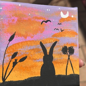 May include: A small, square canvas painting depicting a sunset scene. The artwork features a black silhouette of a rabbit, birds, and plants against a backdrop of orange and purple hues. A crescent moon and stars are also visible.
