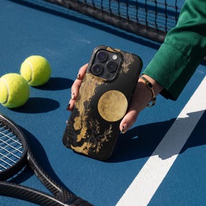 May include: A black smartphone case featuring a gold-colored circular moon graphic and abstract gold-colored swirling patterns against a dark background. The case has a cutout for a triple-lens camera system on the upper left corner.