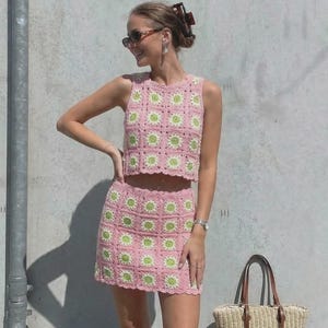 May include: A pink crochet two-piece set featuring a sleeveless top and a mini skirt, both adorned with green floral patterns. A straw tote bag with brown leather handles sits nearby. The outfit is a summer fashion statement.