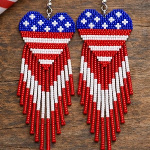 May include: A pair of patriotic earrings featuring a heart-shaped design with a blue beaded top adorned with white stars. The lower portion showcases red and white beaded stripes, creating a fringed effect. The earrings are suspended from silver-colored hooks.