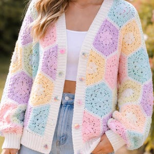 Pode incluir: Um cardigan de crochê em tons pastel com um padrão geométrico. O cardigan apresenta uma mistura de formas hexagonais rosa, amarelas, azuis e lavanda, com acabamentos e botões brancos. As mangas são compridas e o estilo é aberto.