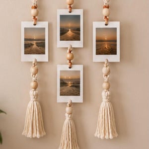 Op de afbeelding: Een wandhanger met een horizontale houten stok en drie verticale macram&eacute;koorden. Elk koord bevat witte rechthoekige fotolijsten met zonsondergang-strandsc&egrave;nes, geaccentueerd met natuurlijke houten kralen en afgewerkt met lange kwastjes.